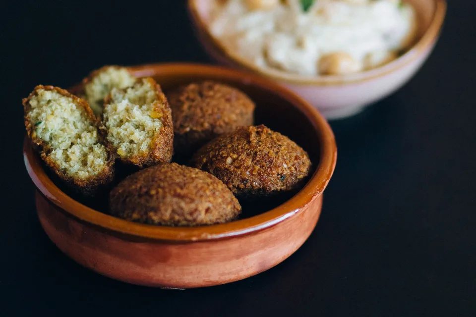 BookNoFees: Fotografia ilustrativa: Bolinhas de falafel, incluindo uma aberta, numa taça com húmus ao fundo